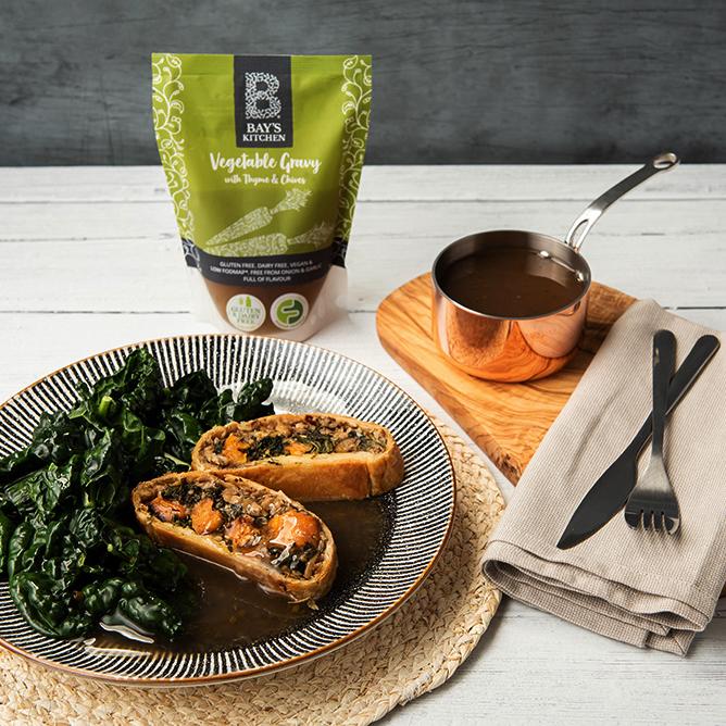 A packet of Bay's Kitchen Vegetable Gravy beside a plate of nut roast Wellington and vegetables