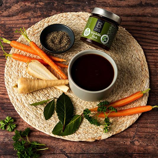 A ramekin of Bay's Kitchen Vegetable stock on a circular wicker placemat with parsnips and carrots